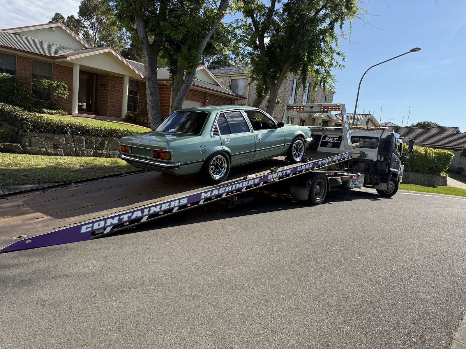 Tilt tray tilted on a suburban street, loading an old green Commodore